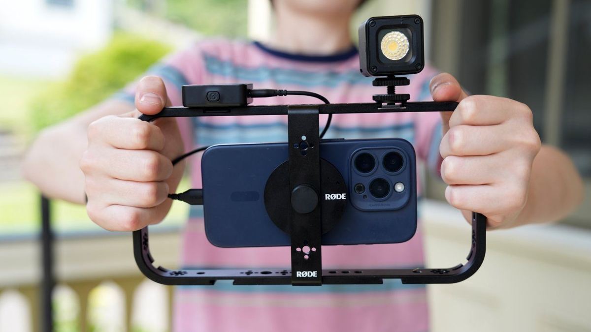Hands holding a smartphone mounted on a stabilizer rig with a Rode microphone and light attached, in an outdoor setting.
